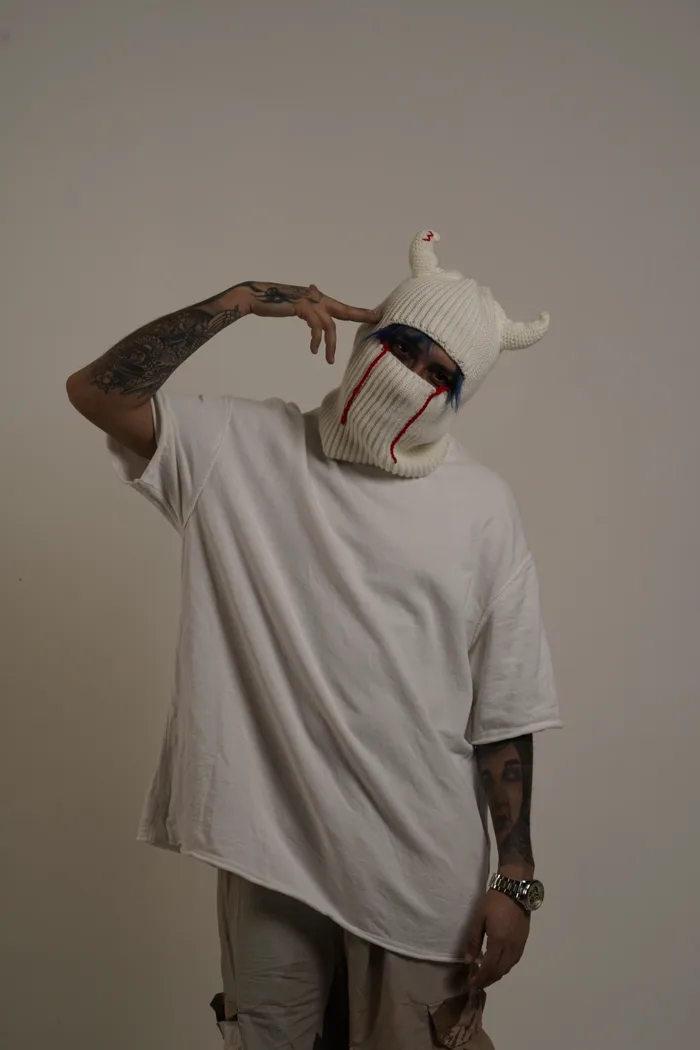 SEVEN77 en cagoule blanche à cornes aux larmes rouges brodées, tee-shirt blanc oversize, touchant une corne de deux doigts