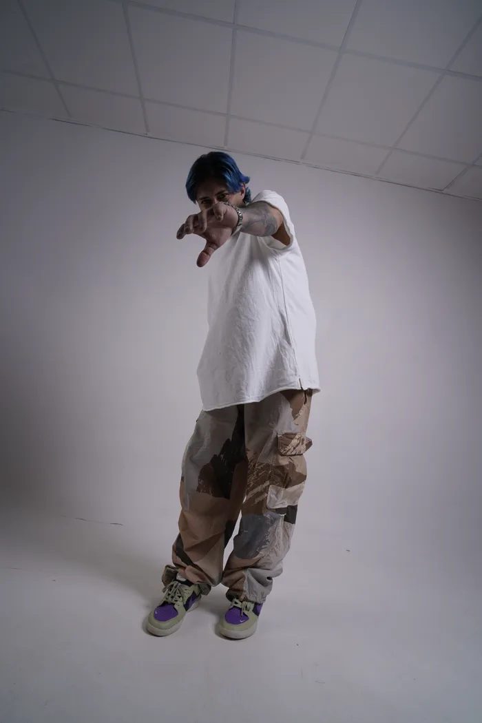 SEVEN77 en pied, cheveux bleus et tee-shirt blanc oversize, pointant la caméra de la main