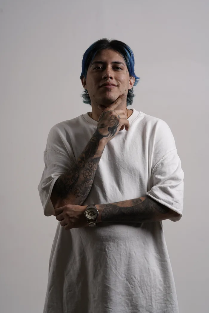 Portrait de SEVEN77 aux cheveux bleus, la main tatouée sous le menton et une montre argentée