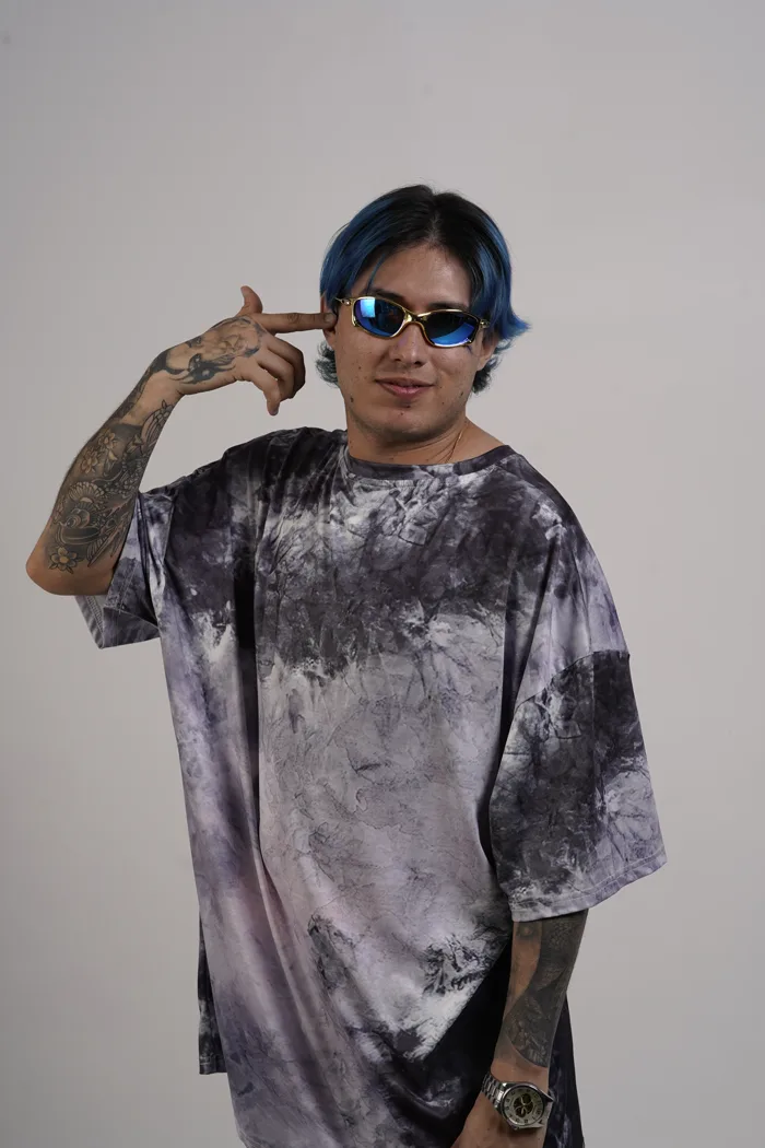SEVEN77 en tee-shirt tie-dye gris et lunettes miroir bleues, mimant un pistolet contre sa tempe