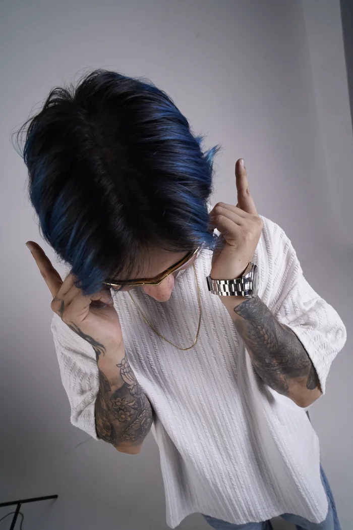 SEVEN77 photographié d'en haut, tête baissée, les mains faisant des cornes près des cheveux bleus